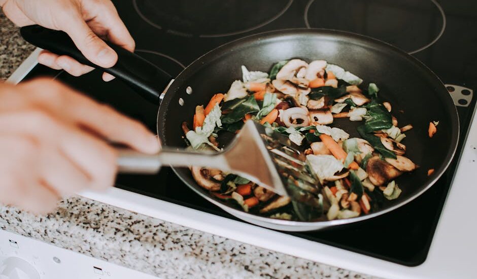 A person stirring vibrant vegetables in a frying pan on an electric stove.