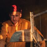 A thoughtful man in a red cap and yellow shirt working in a dimly lit workshop.