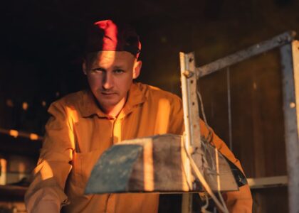 A thoughtful man in a red cap and yellow shirt working in a dimly lit workshop.