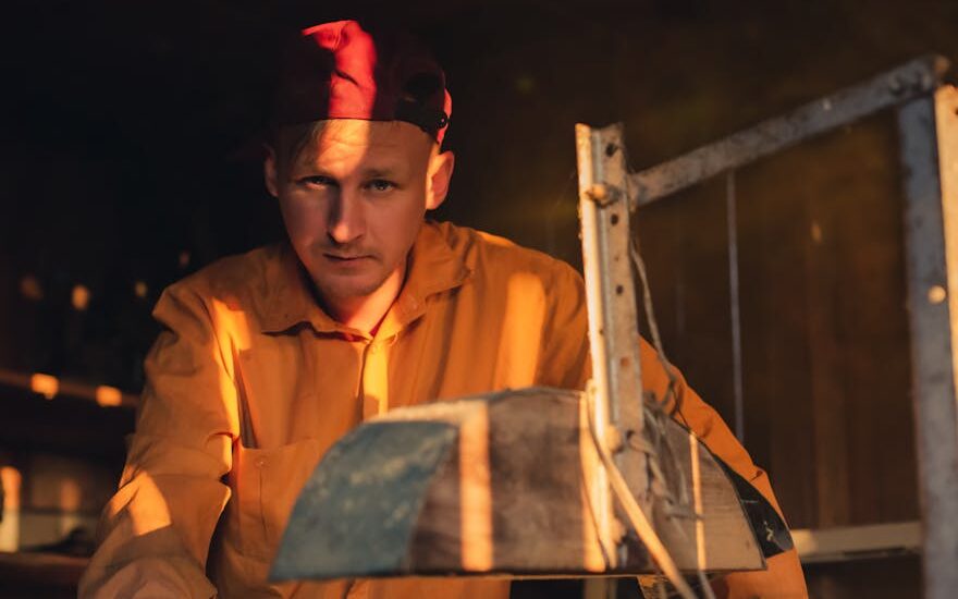 A thoughtful man in a red cap and yellow shirt working in a dimly lit workshop.