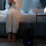 A person in pajamas sits on a bed near a running humidifier, illustrating how to choose a humidifier for bedroom comfort.