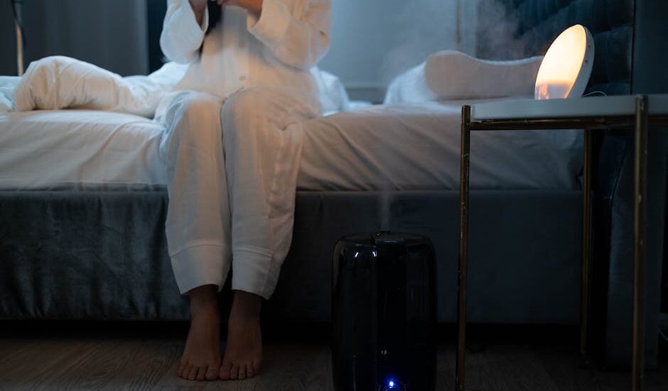 A person in pajamas sits on a bed near a running humidifier, illustrating how to choose a humidifier for bedroom comfort.