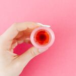 A detailed view of a menstrual cup with red liquid held by a hand against a pink backdrop, symbolizing menstruation.