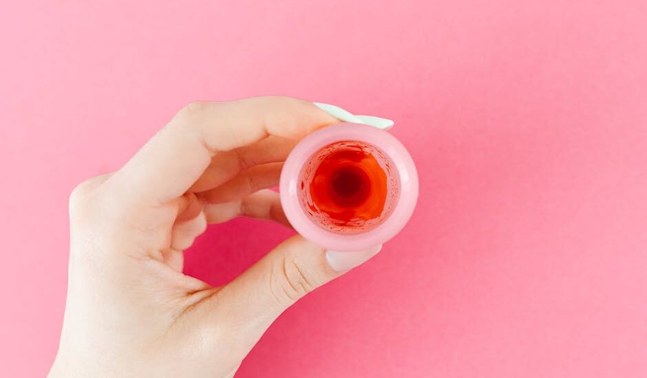 A detailed view of a menstrual cup with red liquid held by a hand against a pink backdrop, symbolizing menstruation.