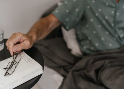 Person in bed reaching for eyeglasses on a bedside table, illustrating a relaxed morning routine while weaning off CPAP therapy.
