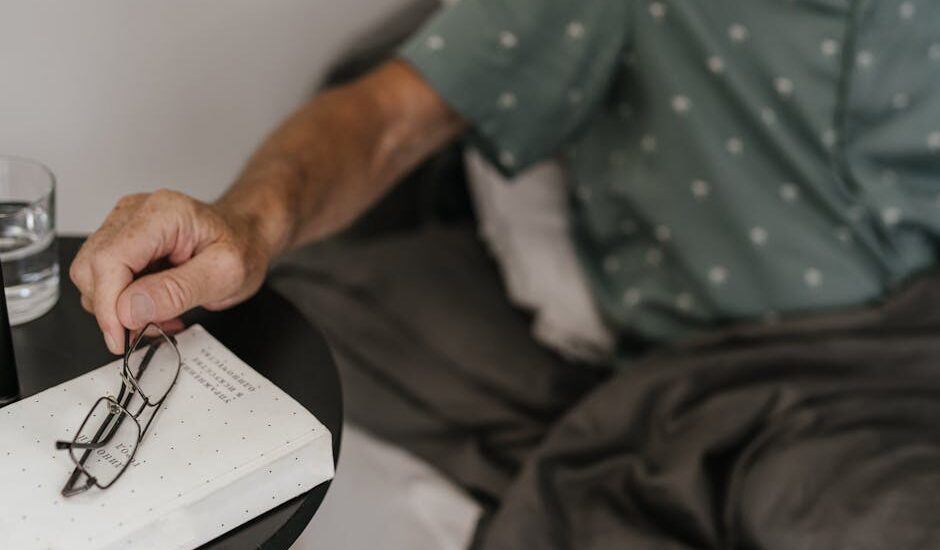 Person in bed reaching for eyeglasses on a bedside table, illustrating a relaxed morning routine while weaning off CPAP therapy.
