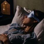 A woman sleeping peacefully wearing a blue eye mask in a cozy, dimly lit bedroom.