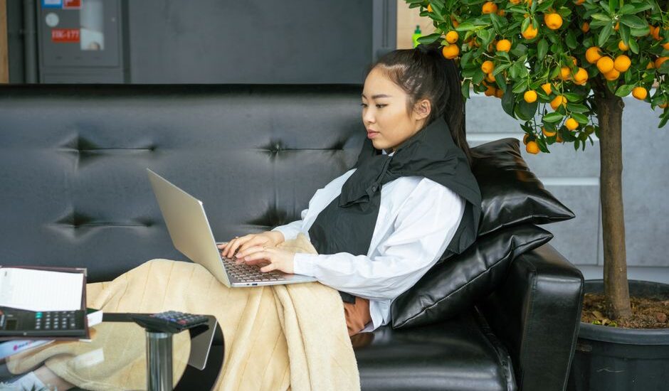 Person relaxing on a black sofa with an infrared heating pad draped over their shoulders, working on a laptop, for an article about infrared heating pads worth
