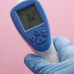 Close-up of an infrared thermometer displaying temperature, held by a gloved hand against a pink background.