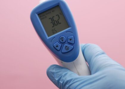 Close-up of an infrared thermometer displaying temperature, held by a gloved hand against a pink background.