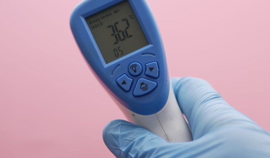 Close-up of an infrared thermometer displaying temperature, held by a gloved hand against a pink background.