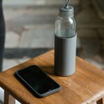 Close-up of a smartphone and water bottle placed on a wooden stool inside a room.