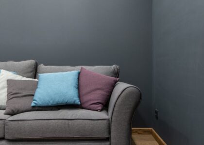 Part of soft modern gray sofa with armrest and abundance of bright pillows placed near wall in light room at home
