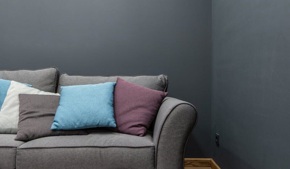 Part of soft modern gray sofa with armrest and abundance of bright pillows placed near wall in light room at home