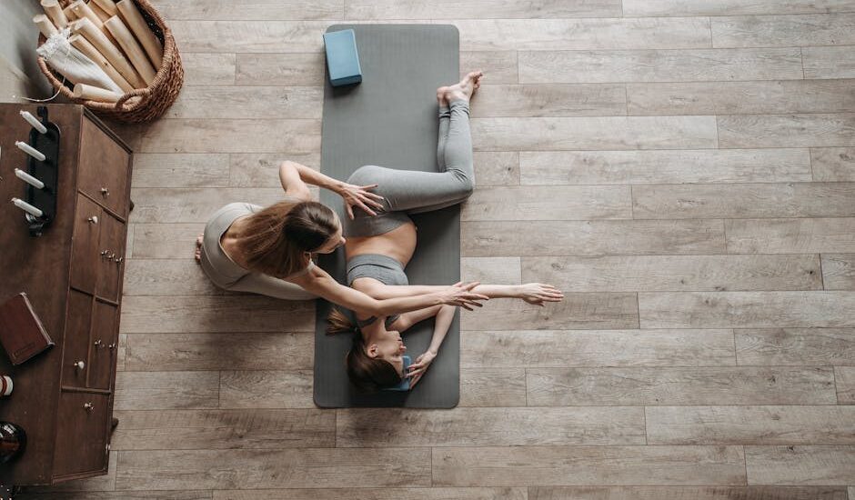 Overhead view of a yoga instructor helping a woman stretch on a mat during an indoor session.