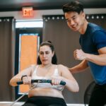 Happy ethnic young instructor supporting plus size woman working out on rowing machine in sport club