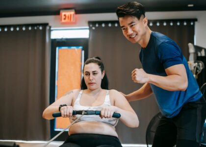 Happy ethnic young instructor supporting plus size woman working out on rowing machine in sport club