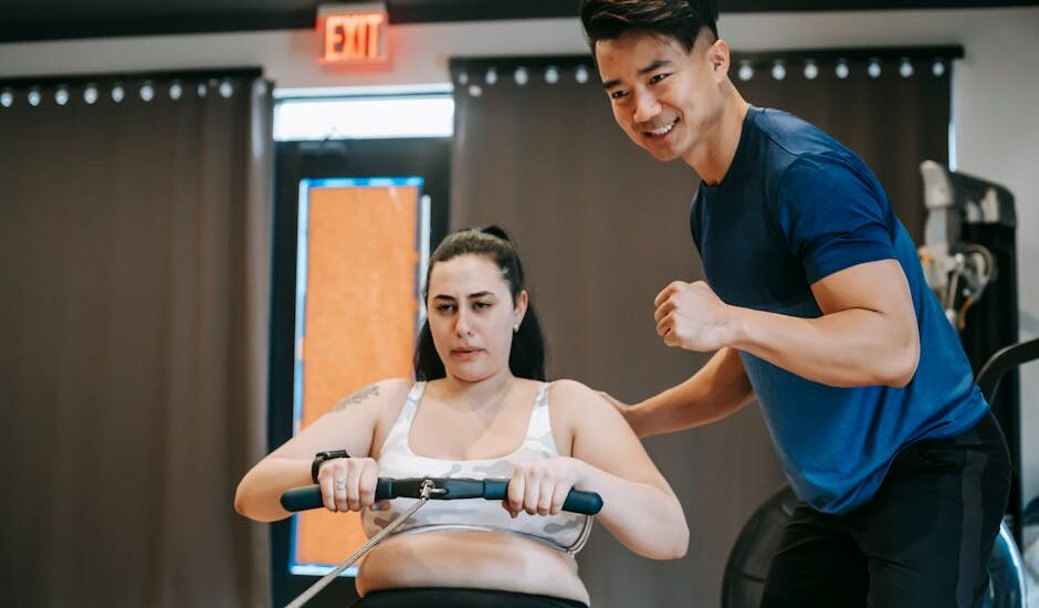 Happy ethnic young instructor supporting plus size woman working out on rowing machine in sport club