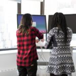 Two people adjust a sit-stand desk converter holding two monitors in a bright office, illustrating dual monitor use.