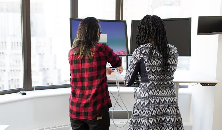 Two people adjust a sit-stand desk converter holding two monitors in a bright office, illustrating dual monitor use.
