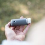 smart inhalers for asthma — Close-up of a hand holding a grey inhaler in a natural outdoor setting.