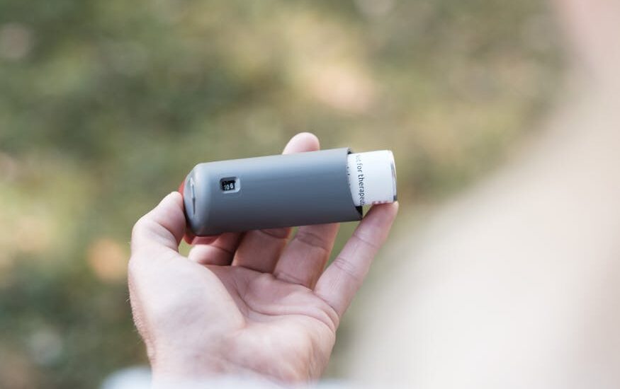 smart inhalers for asthma — Close-up of a hand holding a grey inhaler in a natural outdoor setting.