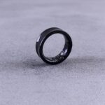 A modern black smart ring with visible circuitry resting on a textured concrete surface.