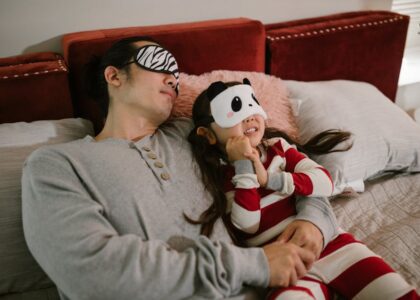 A father and daughter wearing sleep masks relax on a cozy bed, enjoying a moment together.