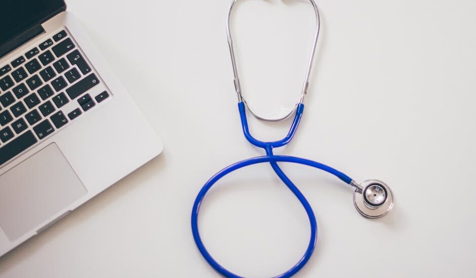 A blue stethoscope and a laptop on a white desk, illustrating the integration of digital stethoscopes in modern healthcare.