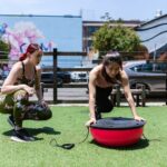 Two women exercising outdoors with a balance board, illustrating fitness equipment options for vibration plate vs balance board comparisons.