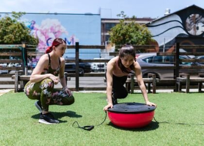 Two women exercising outdoors with a balance board, illustrating fitness equipment options for vibration plate vs balance board comparisons.