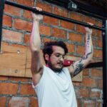 wall mounted pull up bar instructions — A man with tattoos and eyeglasses doing pull-ups inside a gym setting with a brick wall backdrop.