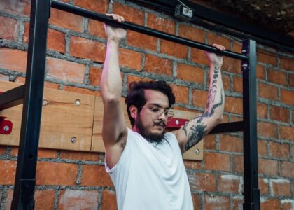 wall mounted pull up bar instructions — A man with tattoos and eyeglasses doing pull-ups inside a gym setting with a brick wall backdrop.
