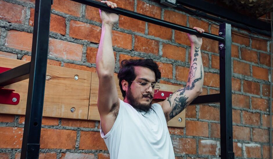 wall mounted pull up bar instructions — A man with tattoos and eyeglasses doing pull-ups inside a gym setting with a brick wall backdrop.