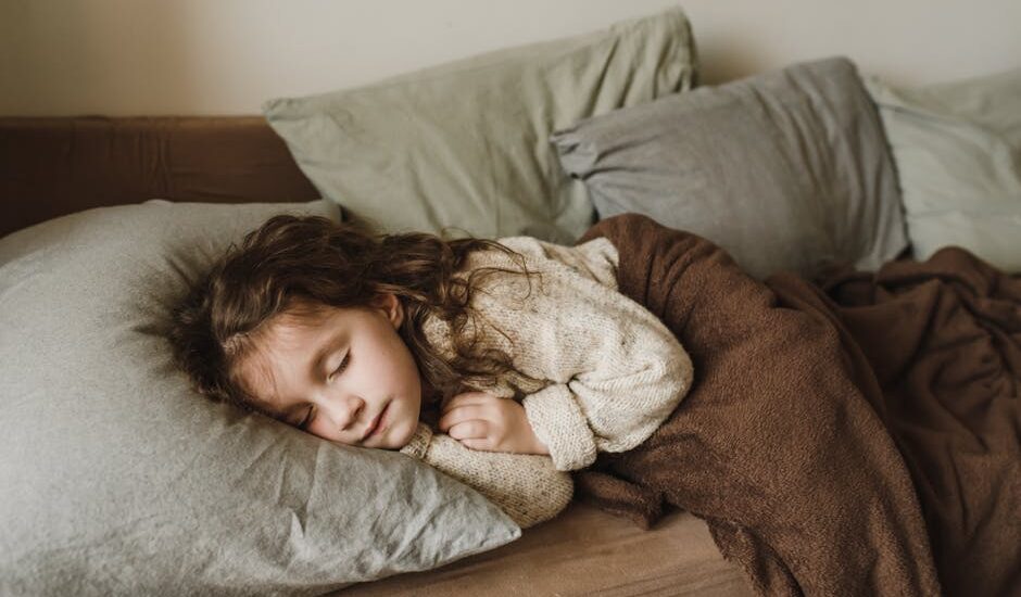 Young child sleeping under a brown blanket on a bed, illustrating considerations for parents choosing weighted blankets for kids.