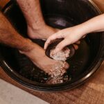 Person soaking feet in a black basin while another hand pours salt crystals, illustrating foot care methods related to foot pads.
