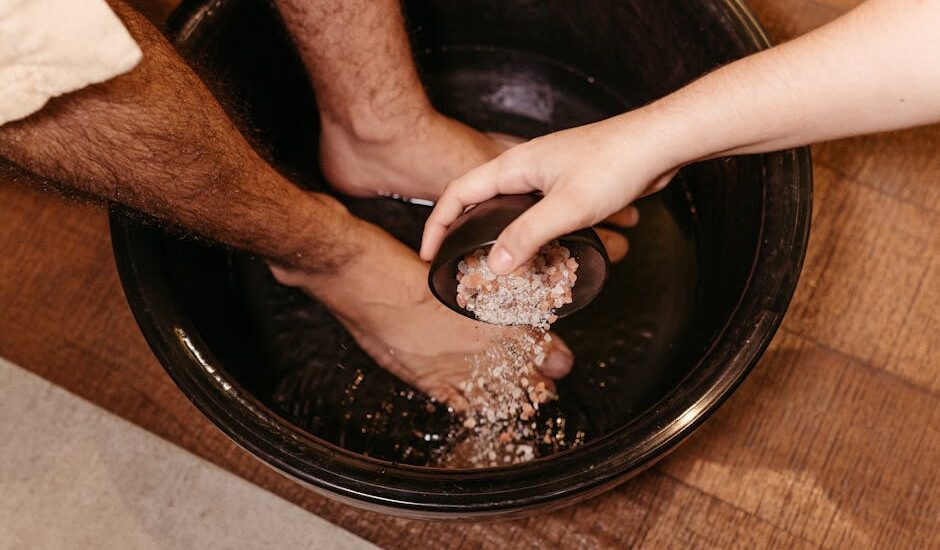 Person soaking feet in a black basin while another hand pours salt crystals, illustrating foot care methods related to foot pads.