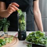 Person preparing a fresh green smoothie with spinach, cucumber, and herbs using a blender.