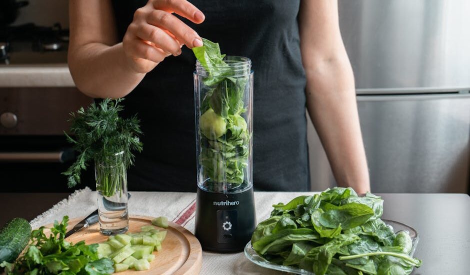 Person preparing a fresh green smoothie with spinach, cucumber, and herbs using a blender.