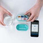 A person using an electronic medication dispenser with a smartphone app and a glass of water.
