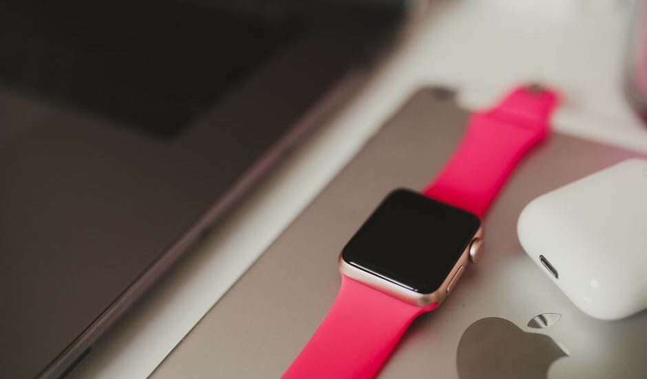A fitness tracker with a pink band is placed next to a laptop and earbuds, illustrating how to charge a fitness tracker.
