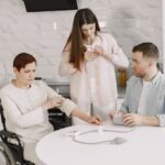 A woman in a wheelchair receives assistance with a portable oxygen concentrator refill in a kitchen.