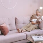 A woman reading on a couch while wearing a heated neck and shoulder wrap for pain relief.