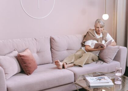 A woman reading on a couch while wearing a heated neck and shoulder wrap for pain relief.
