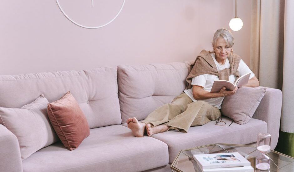 A woman reading on a couch while wearing a heated neck and shoulder wrap for pain relief.